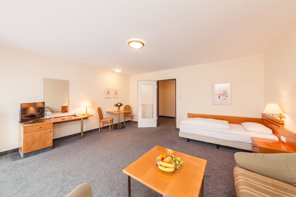 A hotel room with a bed, a desk with a computer, a chair, a small table with fruit, and a sofa.