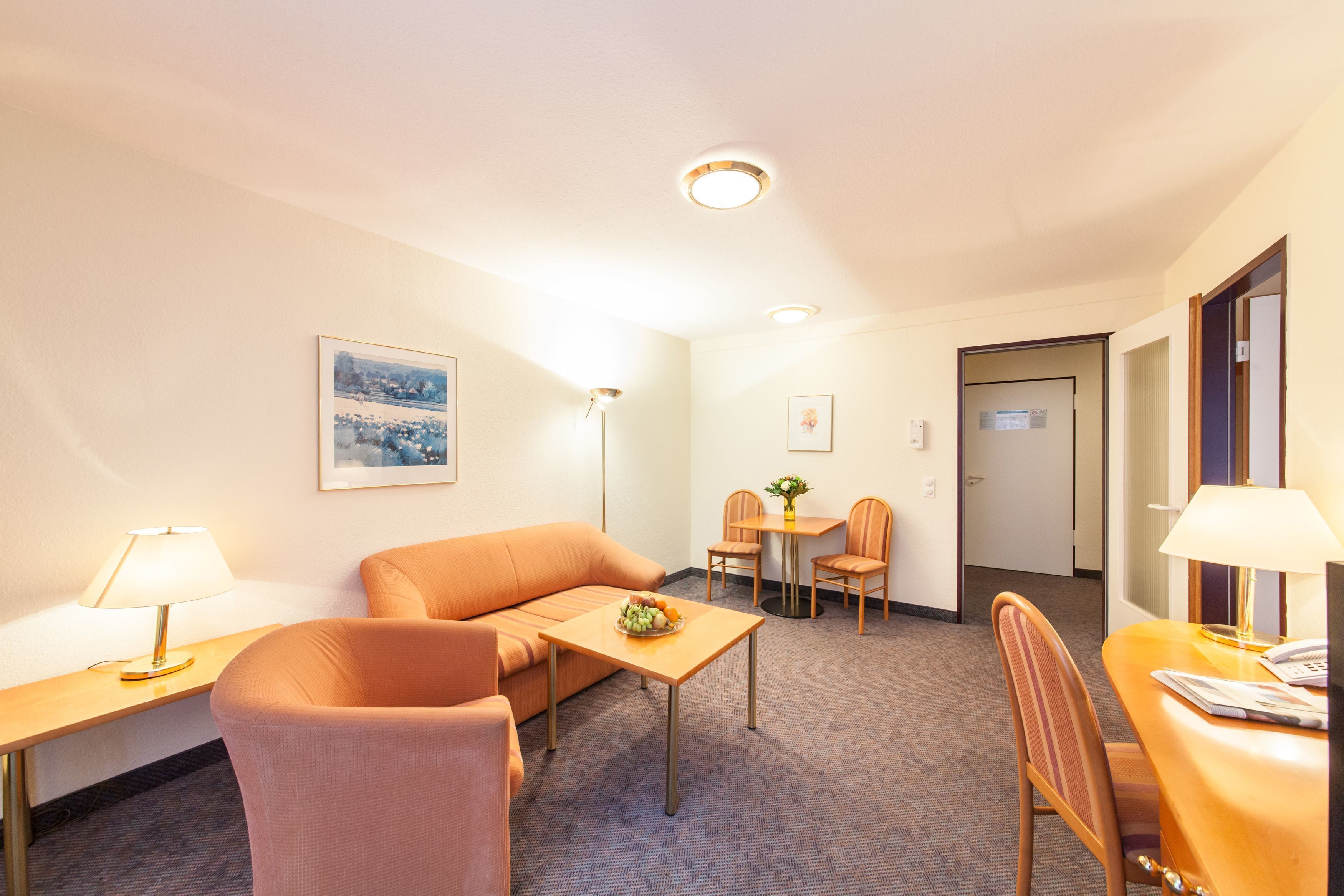A hotel room with a sofa, a small table with fruit, a desk with a lamp, a dining area with chairs, and a painting on the wall.
