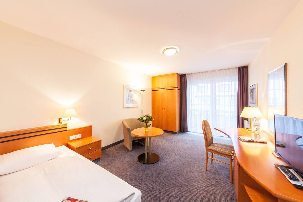 A hotel room with a bed, a desk with a computer, a chair, a small table with a lamp, and a wardrobe.