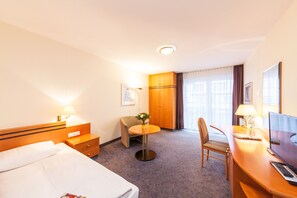 A hotel room with a bed, a desk with a computer, a chair, a small table with a lamp, and a wardrobe. - acora Leipzig Living the City (Leipzig)