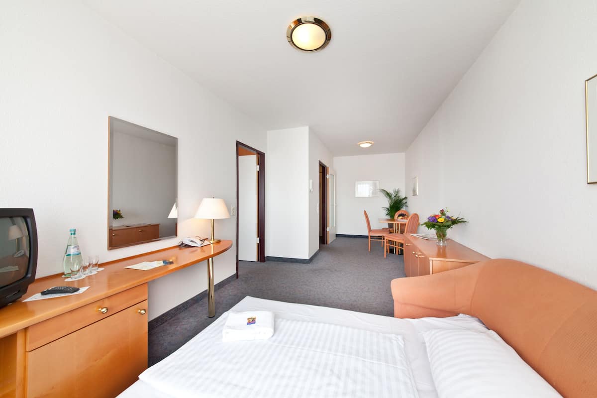 A hotel room with a bed, a desk with a lamp, a television, a mirror, and a small table with a vase of flowers.
