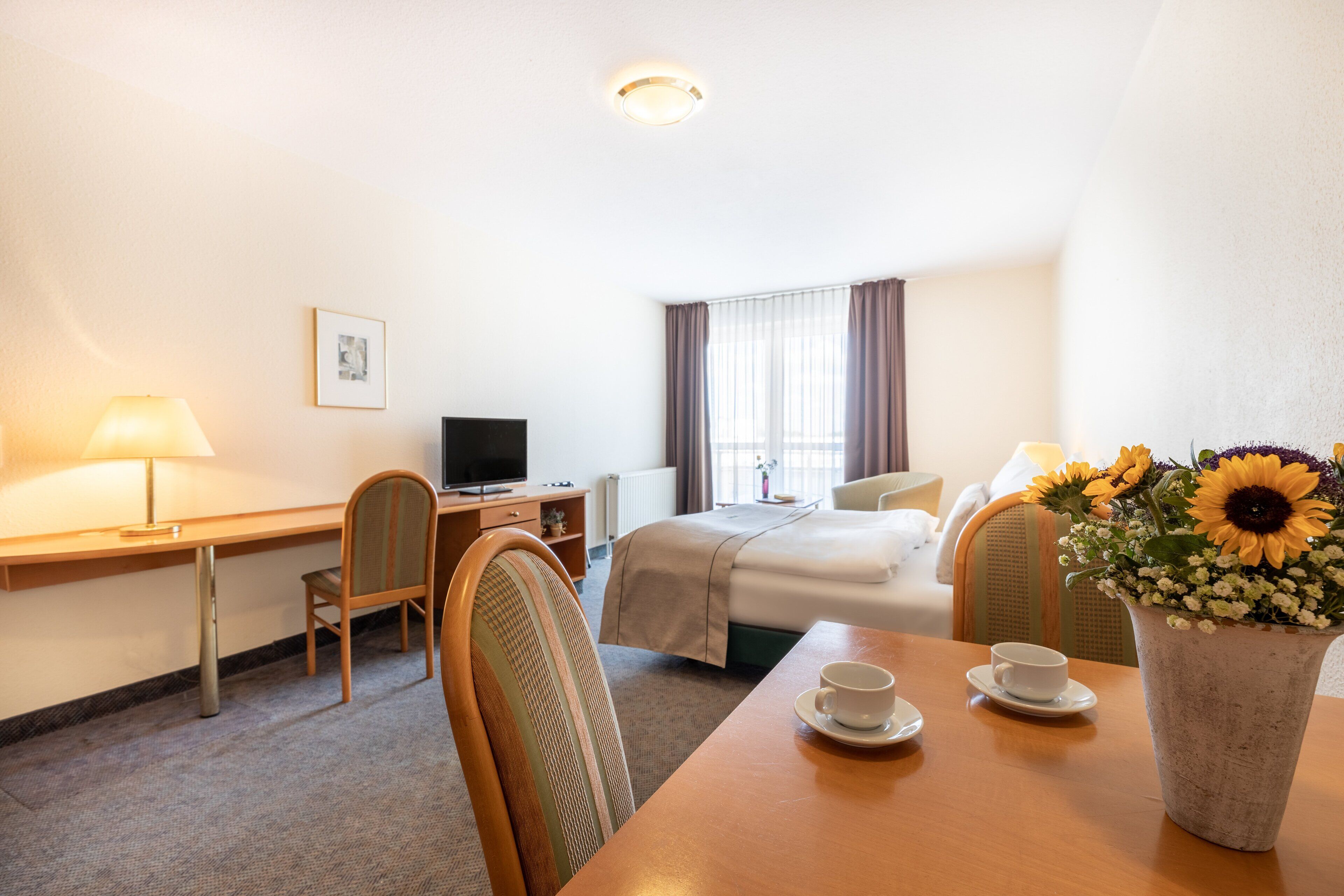 Una camera d'albergo con un letto, una scrivania, una sedia, una TV e un tavolo con un vaso di fiori.