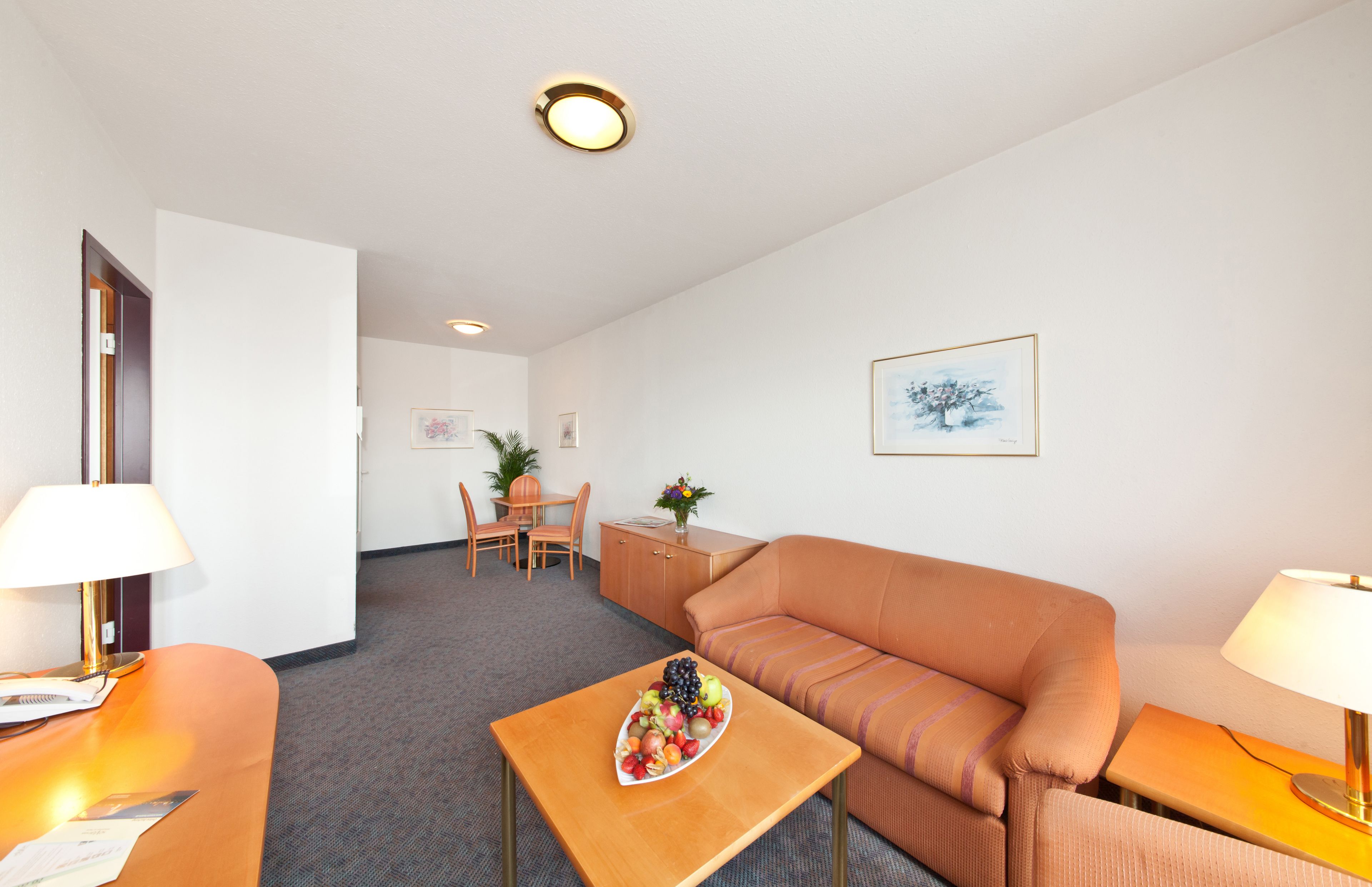 Ein Hotelzimmer mit einer Couch, einem Esstisch mit Obstschale, einer kleinen Kommode und einem Bild an der Wand.