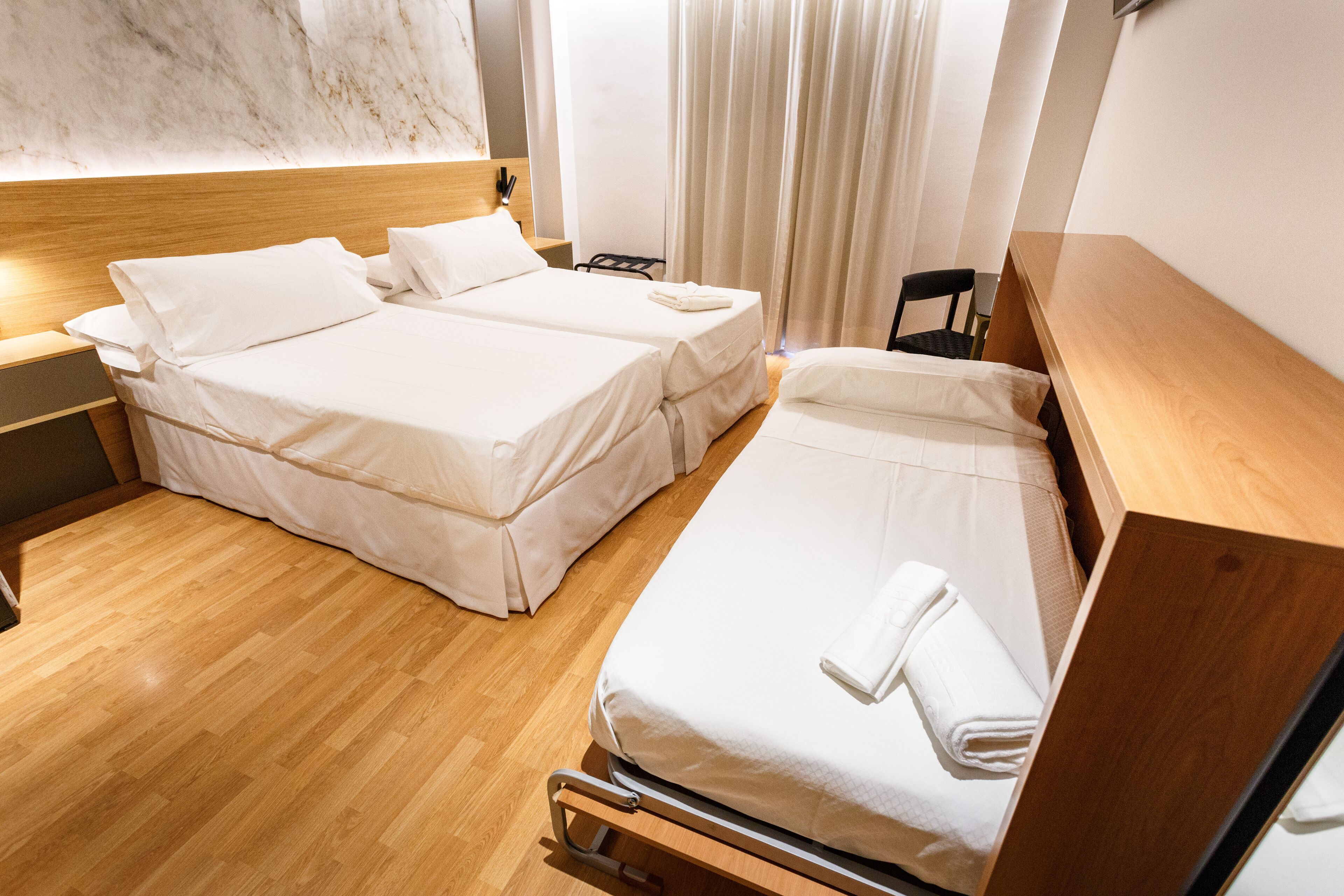 Habitación de hotel con dos camas individuales, suelo de madera, cabecero de madera, una mesita de noche pequeña y una silla.