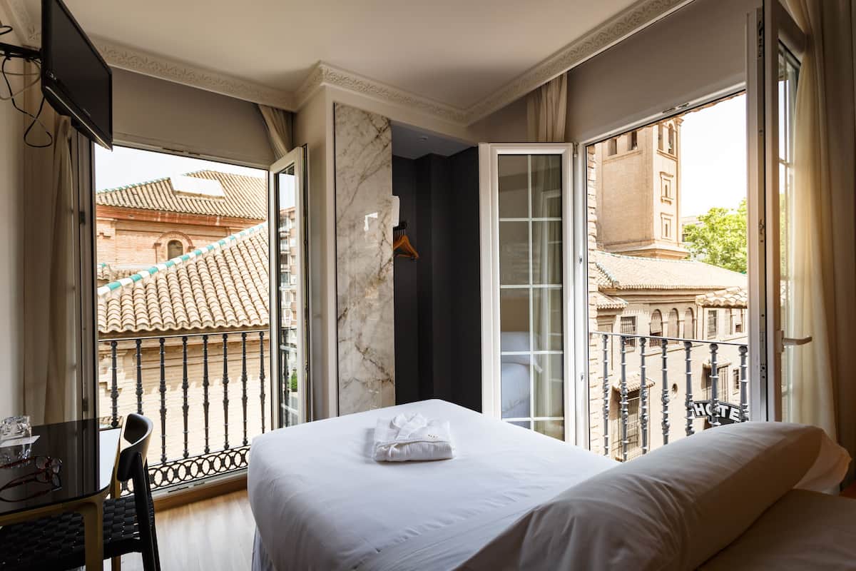 A hotel room with a bed, a desk, and a chair. There is a window with a view of buildings outside.