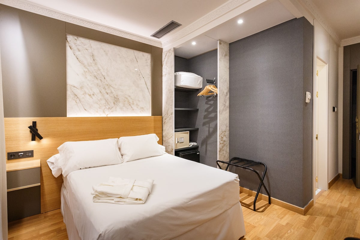 A hotel room with a large bed, a wooden headboard, a marble accent wall, a small desk, a wardrobe, and a chair.