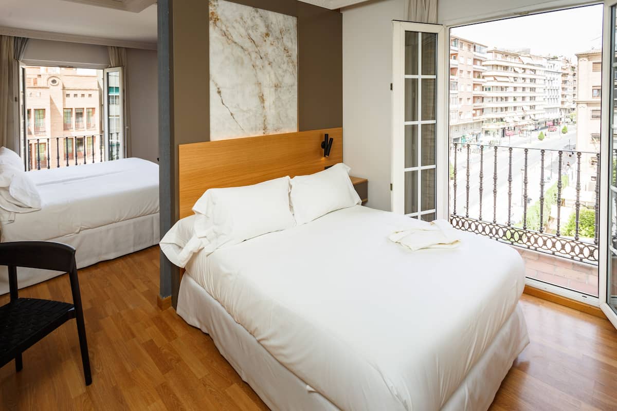 A hotel room with a large bed, a smaller bed, a wooden chair, and a balcony with a view of the city.