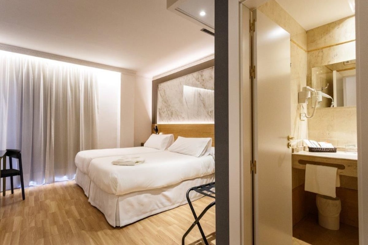 A hotel room with a double bed, a wooden floor, a bathroom with a marble vanity, and a large window with curtains.