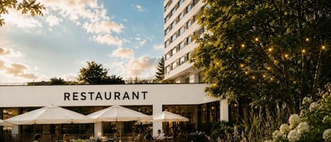 An outdoor restaurant area with tables and umbrellas, surrounded by greenery and a building with a sign that reads 'RESTAURANT'.