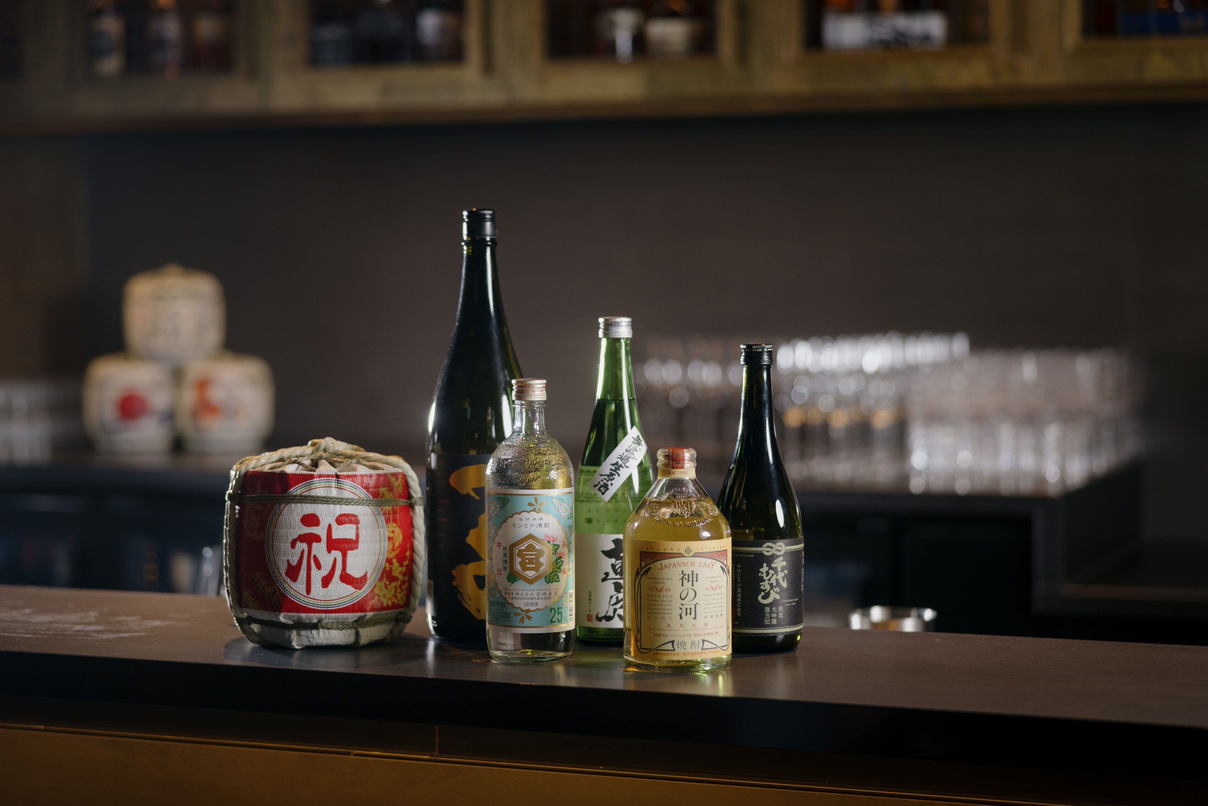 On voit une variété de bouteilles sur le comptoir d’un bar, dont une bouteille de saké japonais traditionnel et plusieurs autres bouteilles portant des étiquettes.