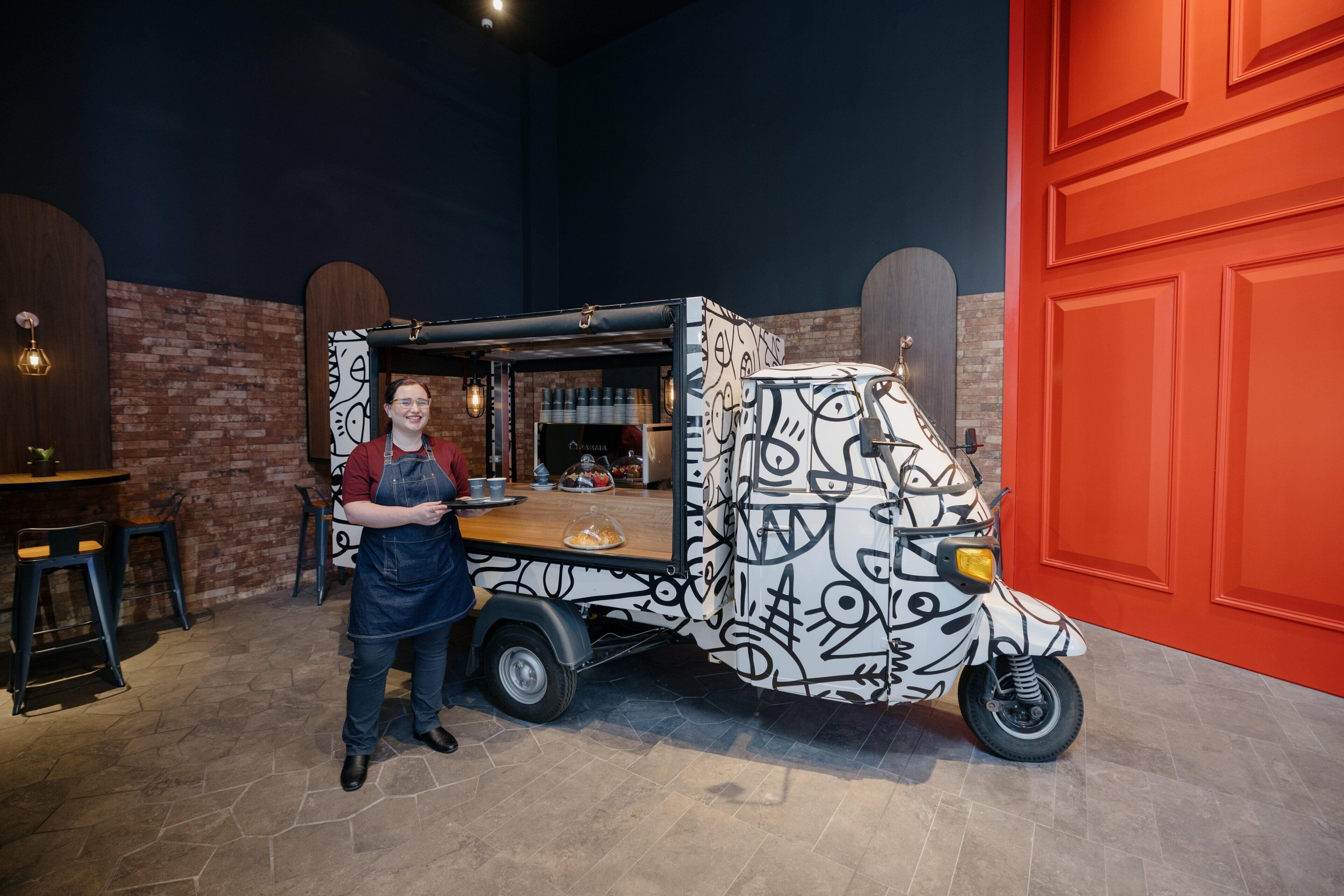 A person standing next to a food truck with a unique graffiti design, a brick wall, and a red door in the background.