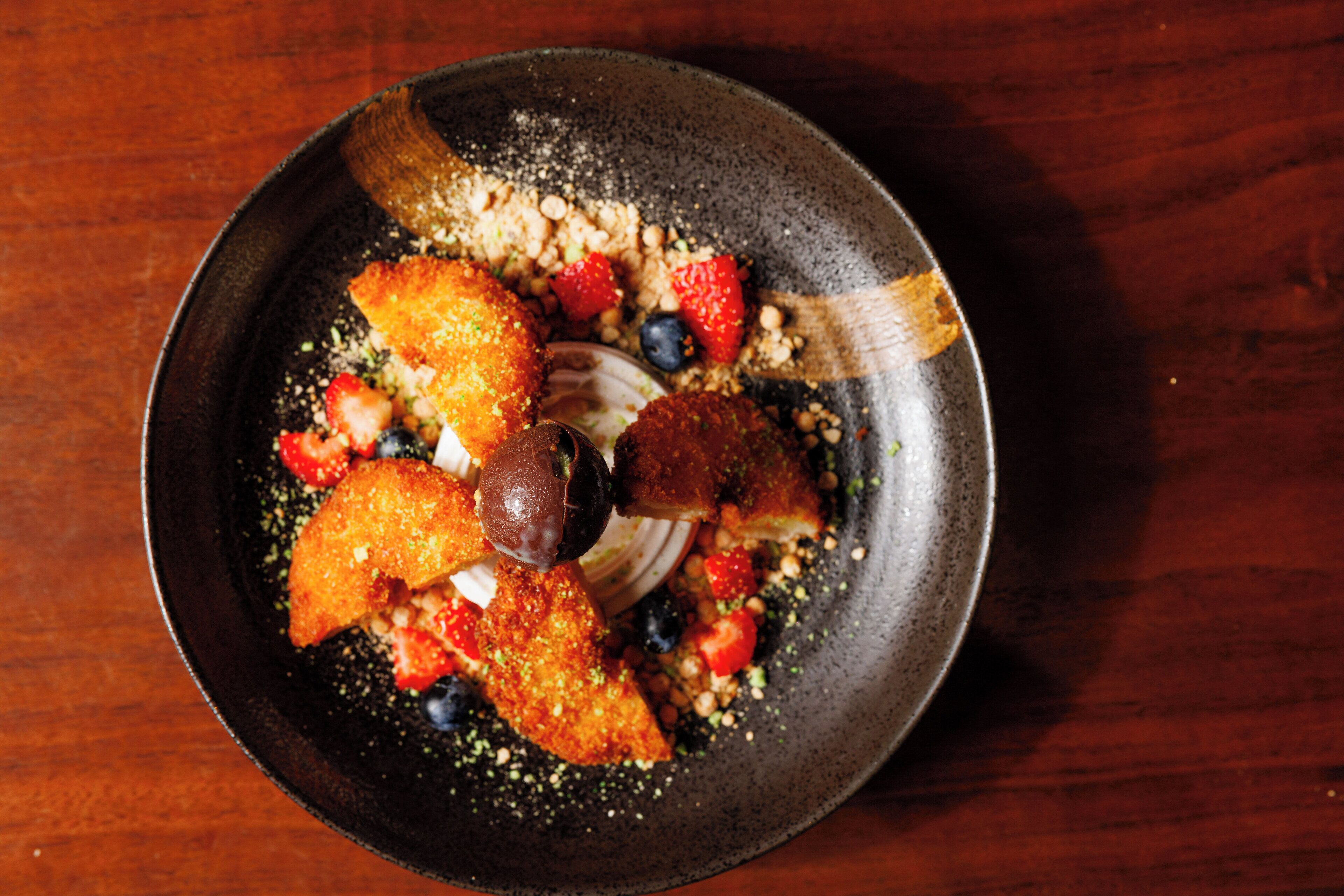 A round, dark-colored plate with a variety of food items including breaded items, strawberries, blueberries, and a dollop of cream.