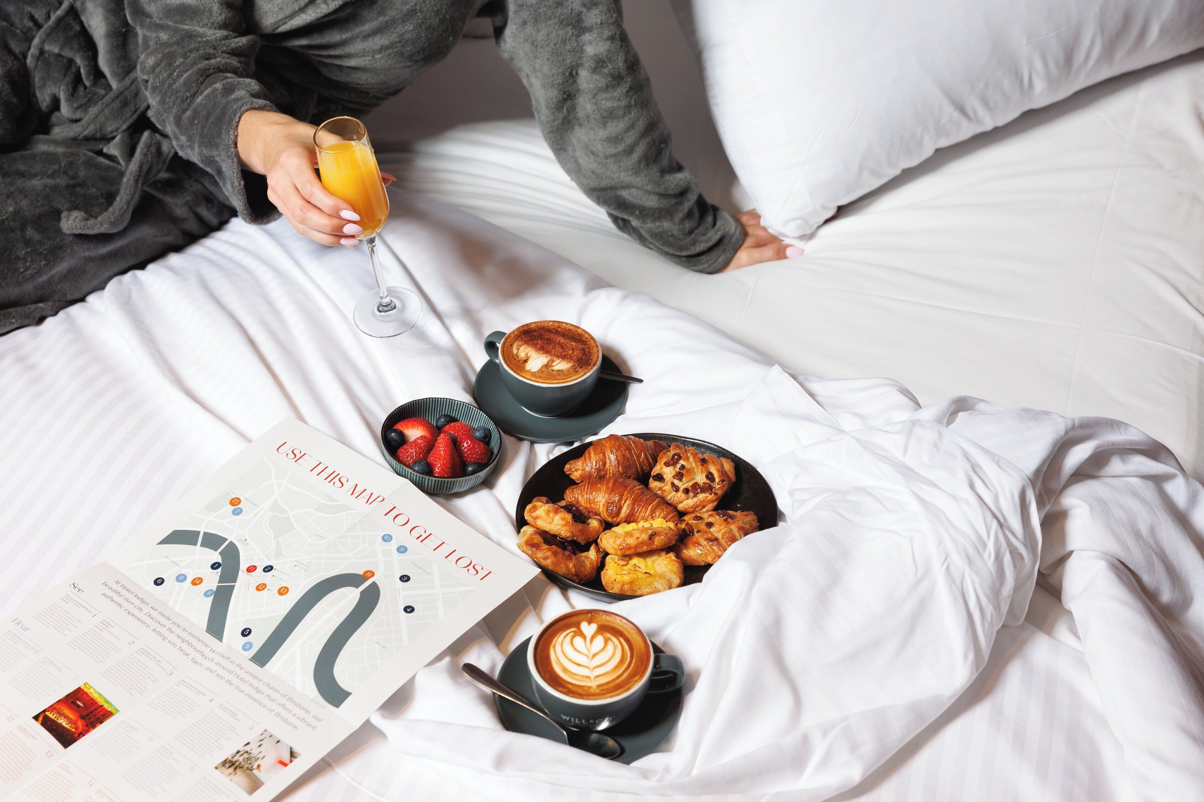 Ein Bett mit weißen Leinen, ein Buch mit dem Titel "USI TEGILLOS", ein Glas Orangensaft, eine Schale Erdbeeren, ein Teller Croissants und zwei Tassen Kaffee.