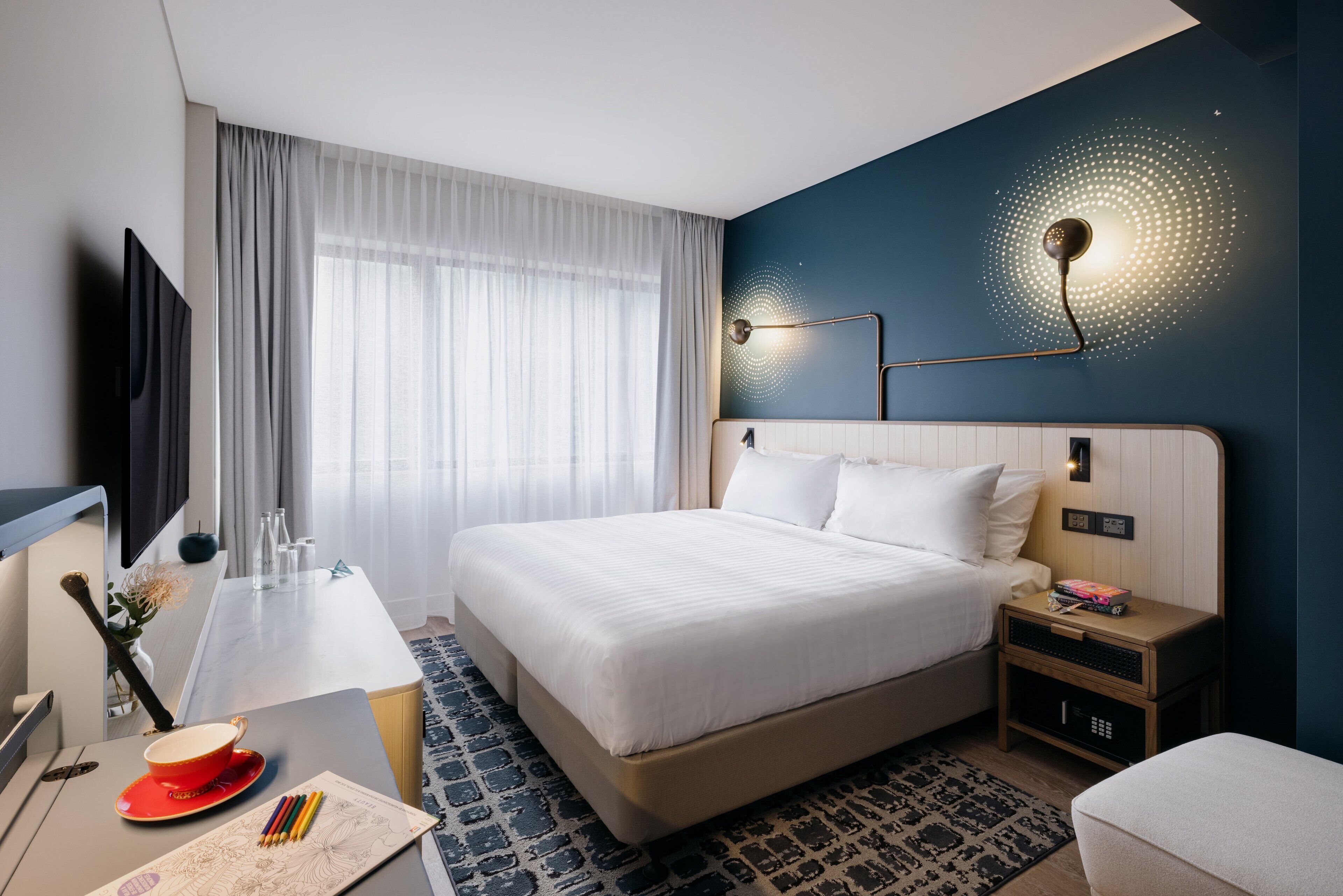 A hotel room with a large bed, a desk with a chair, a TV, and a wall-mounted lamp.