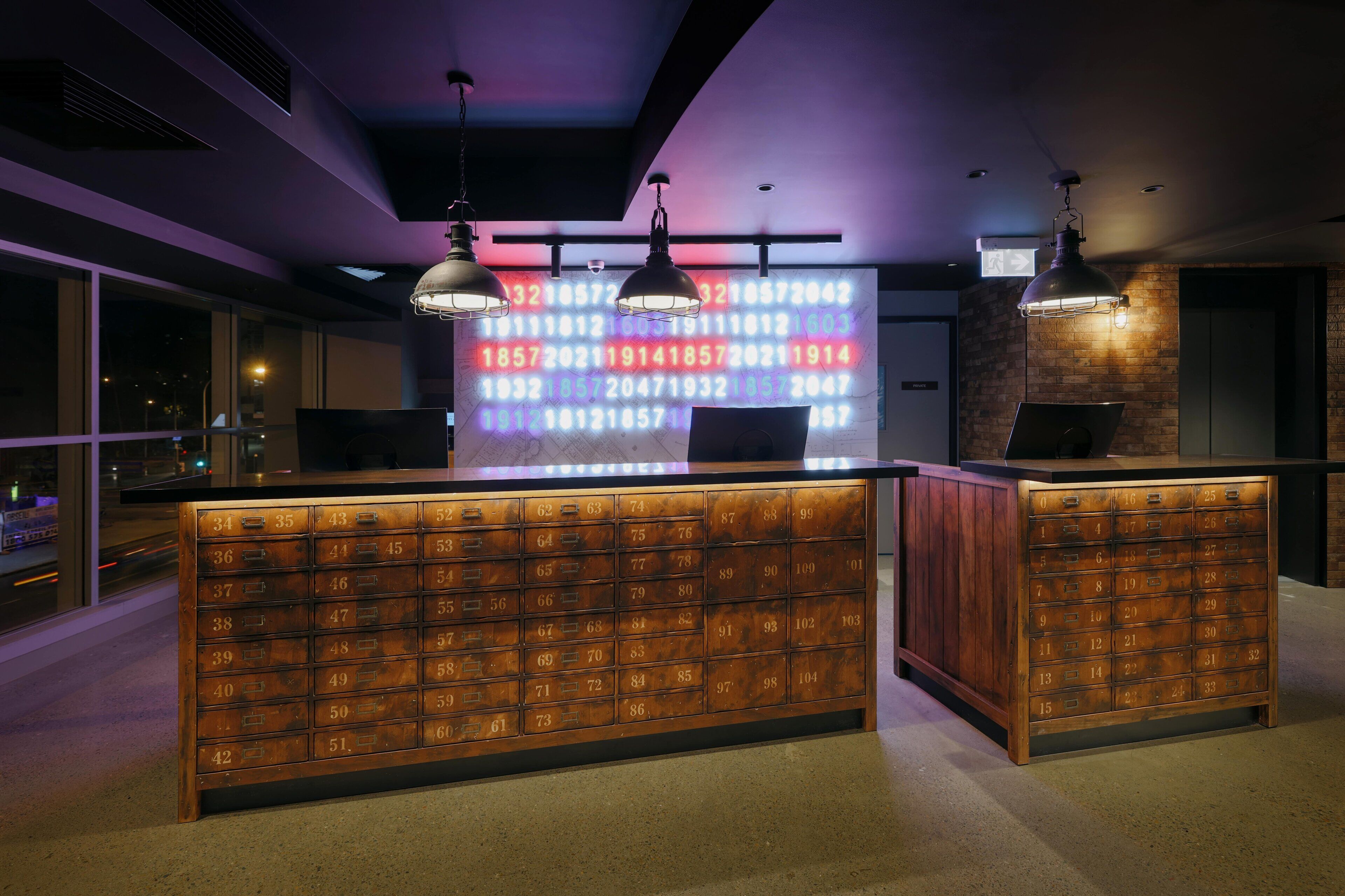 A hotel reception desk with a wooden number lock system, pendant lighting, and a digital display wall.