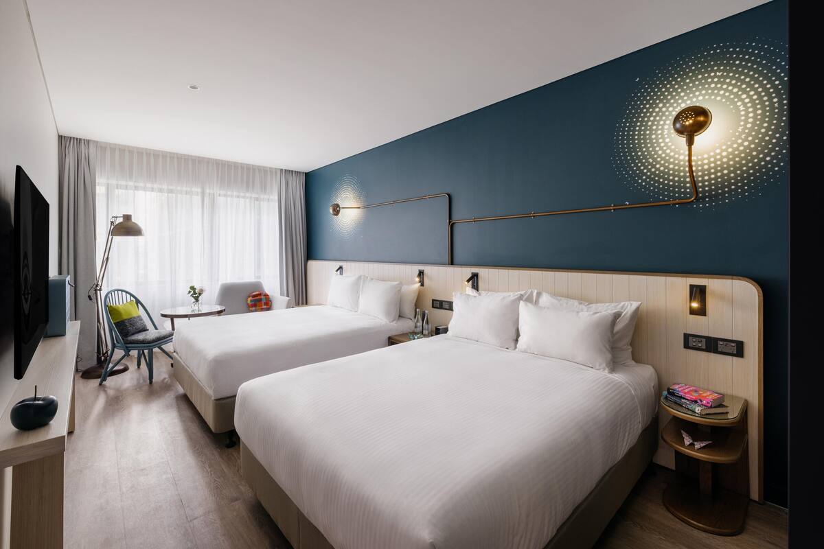 A hotel room with two beds, a desk, a chair, a lamp, and a wall-mounted light fixture.