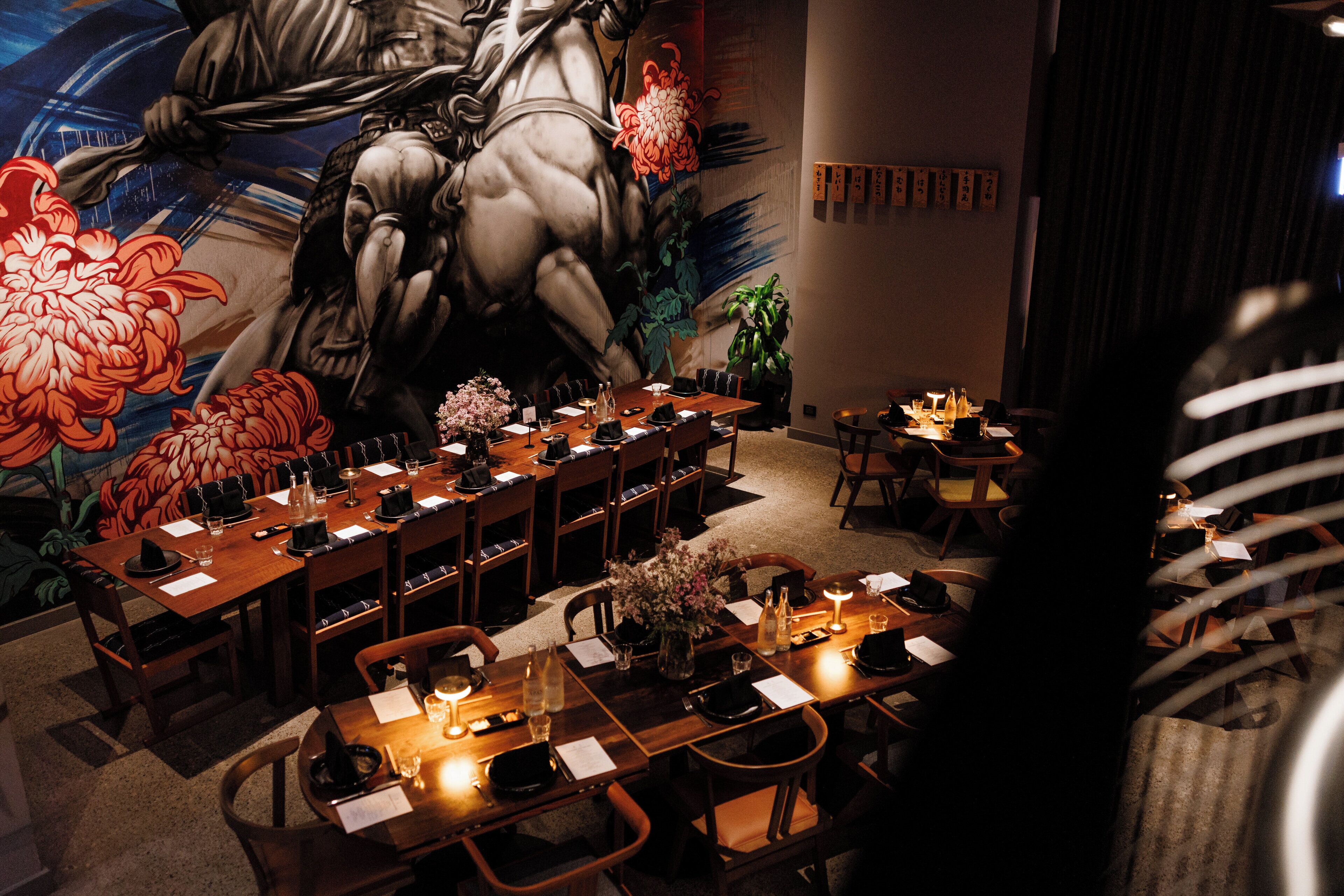 Een restaurant met lange houten tafels gedekt voor een maaltijd, een muurschildering en een potplant.