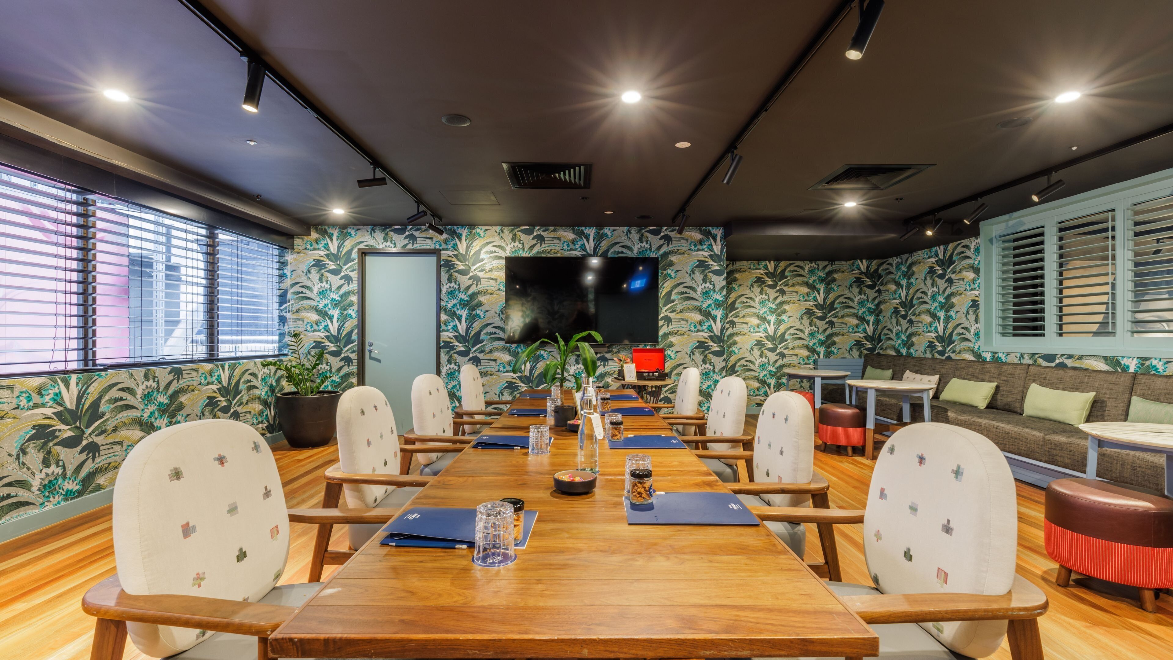 A conference room with a long wooden table, white chairs, a large flat-screen TV, and a patterned wall.