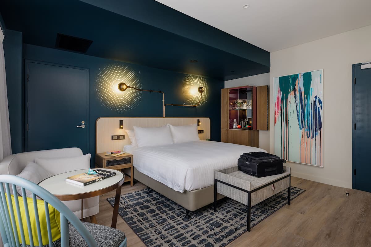 A modern hotel room with a bed, a desk, a chair, a small table, a lamp, a bookshelf, and a large abstract painting.