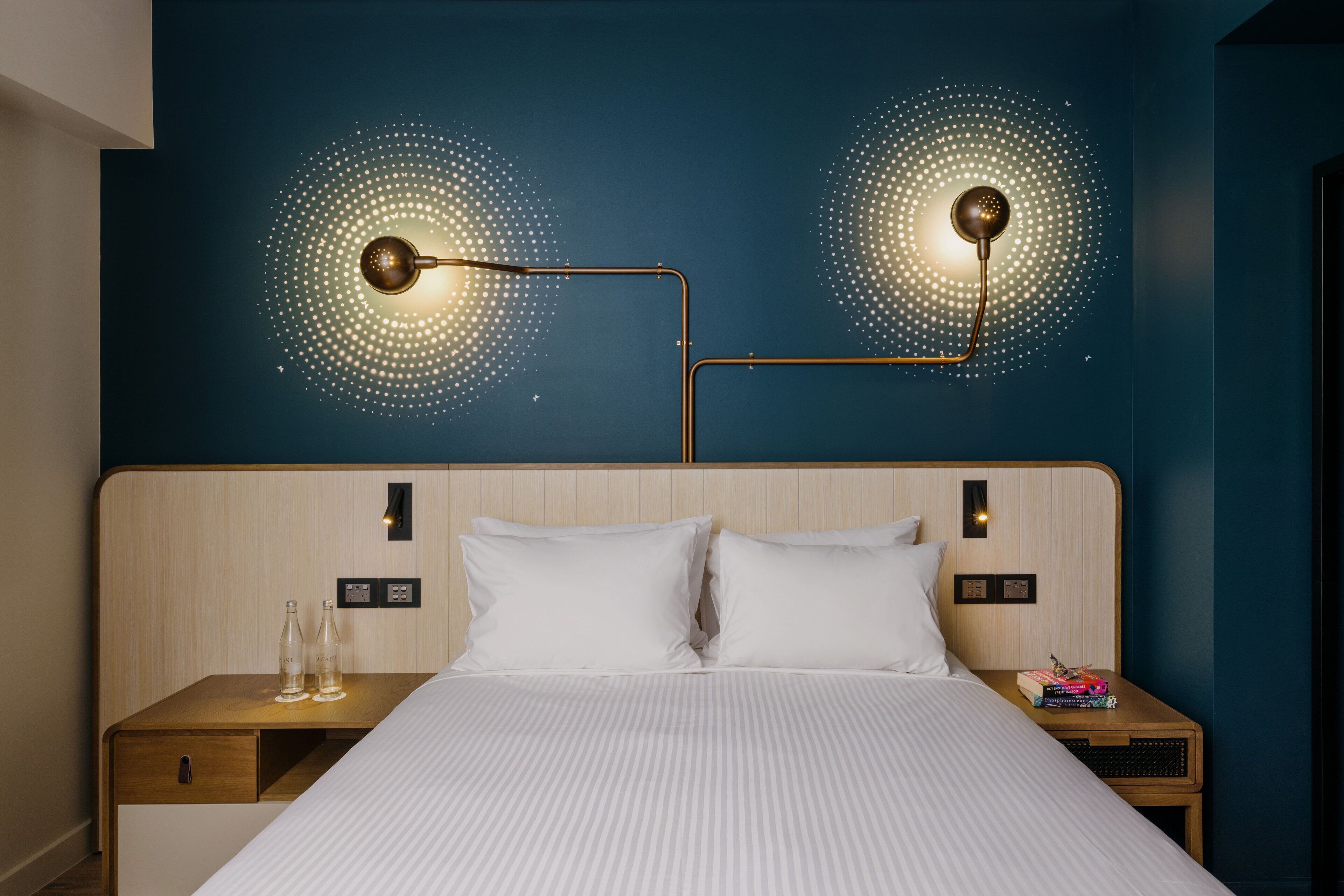 Habitación de hotel con cabecera de madera, dos mesitas de noche, cama con ropa blanca y dos lámparas de pared con un diseño de puntos.