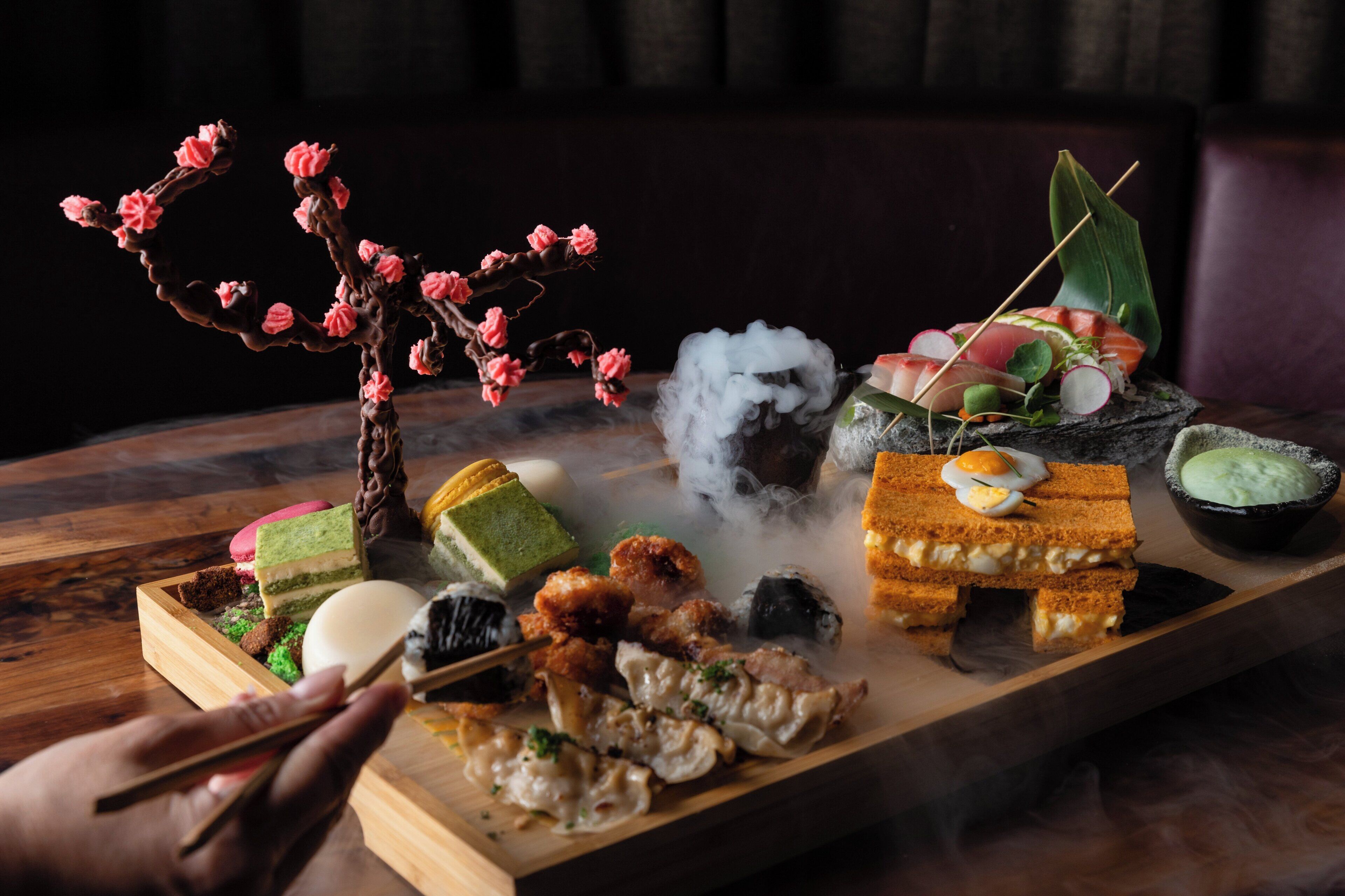 A variety of food items on a wooden tray, including sushi, a dessert, and a decorative branch.