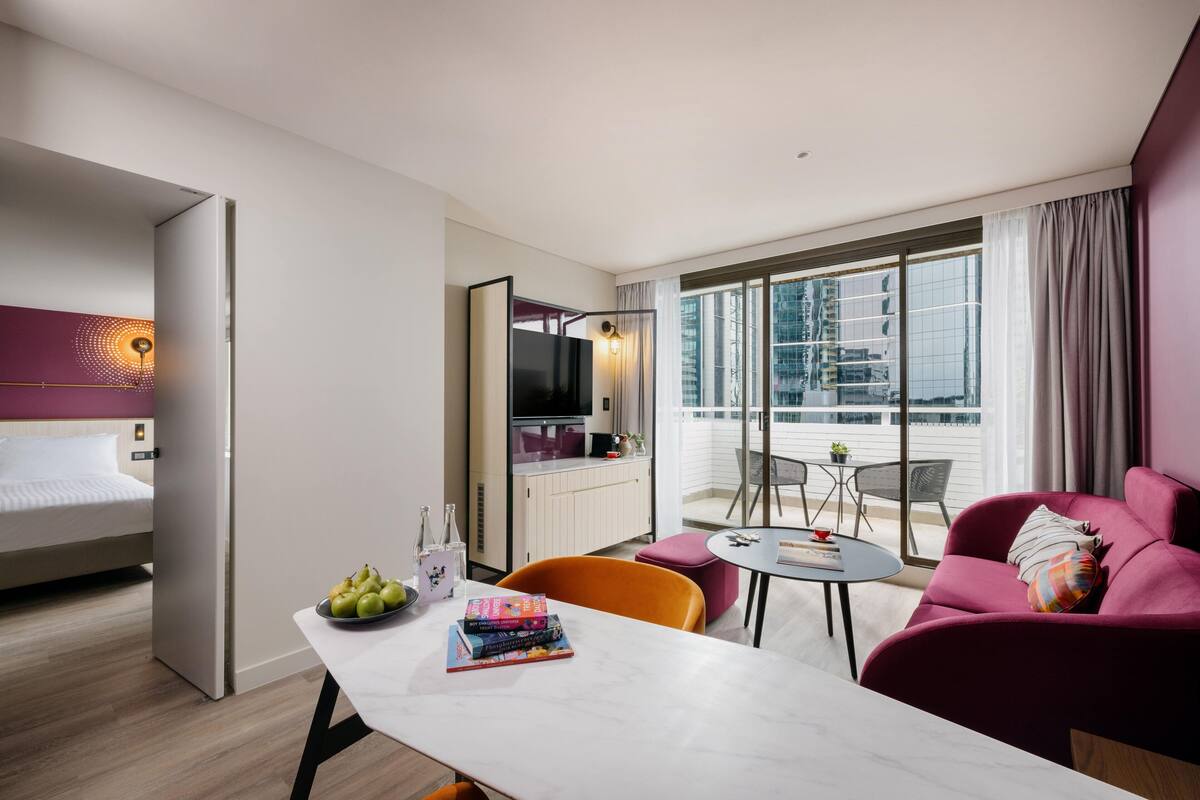 A modern hotel room with a bed, a dining area, a sofa, and a balcony with a city view.
