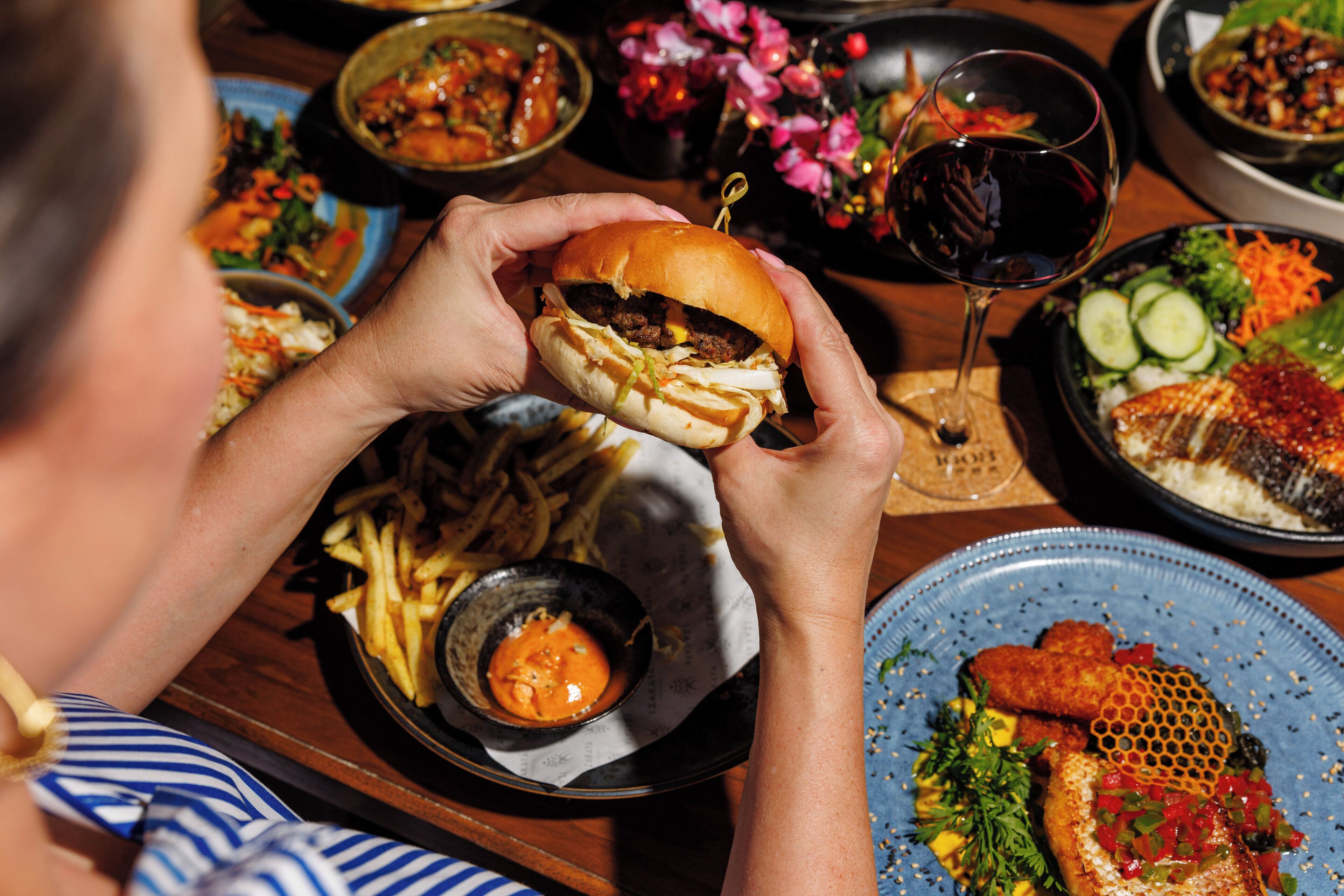 A person holding a burger with fries and a drink on a table with various dishes.