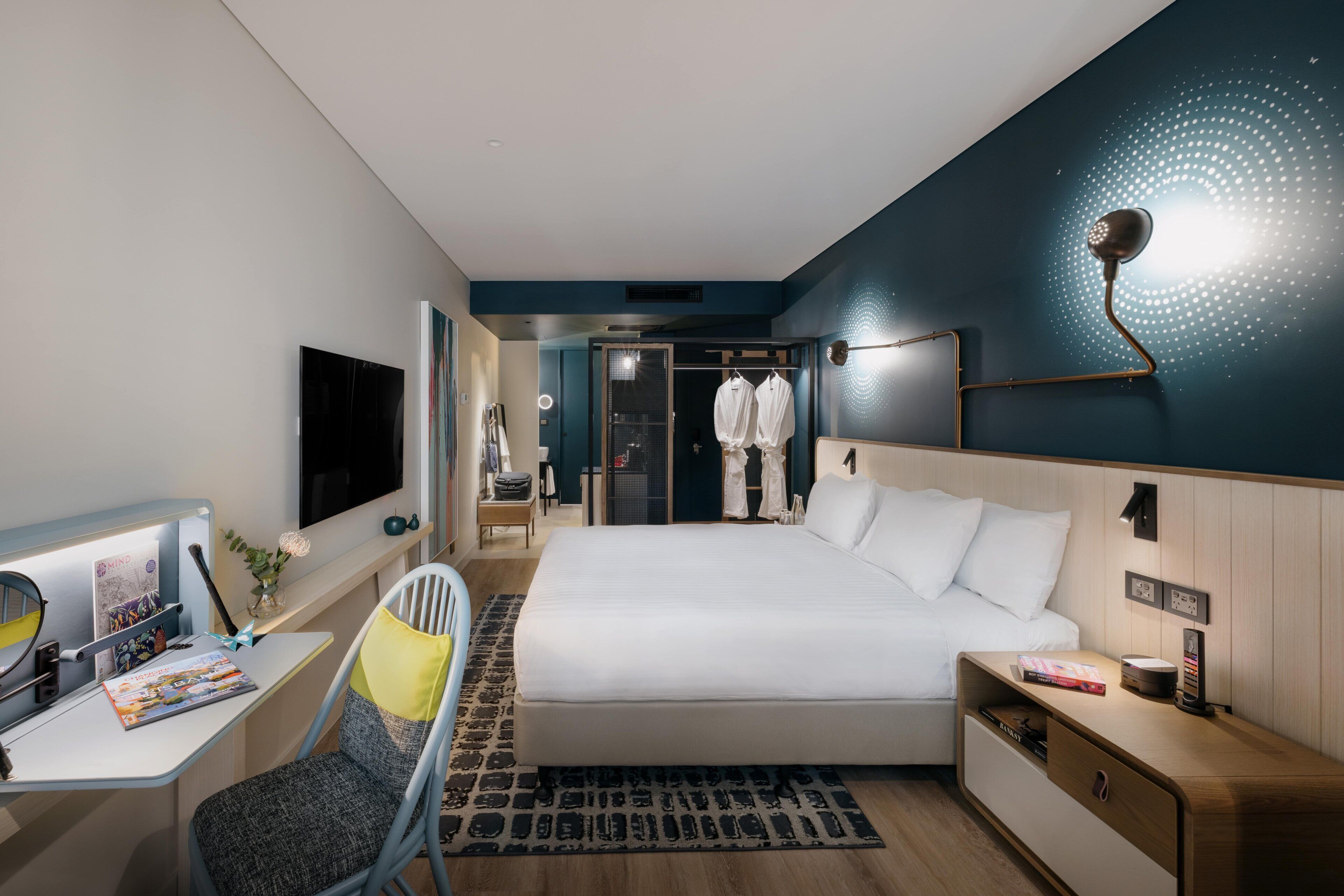 Quarto de hotel moderno com uma cama grande, escrivaninha com cadeira, televisão e guarda-roupa.