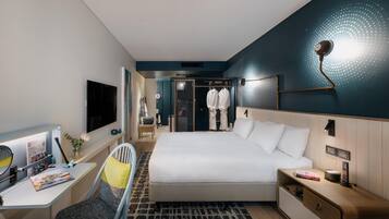 Quarto de hotel moderno com uma cama grande, escrivaninha com cadeira, televisão e guarda-roupa.