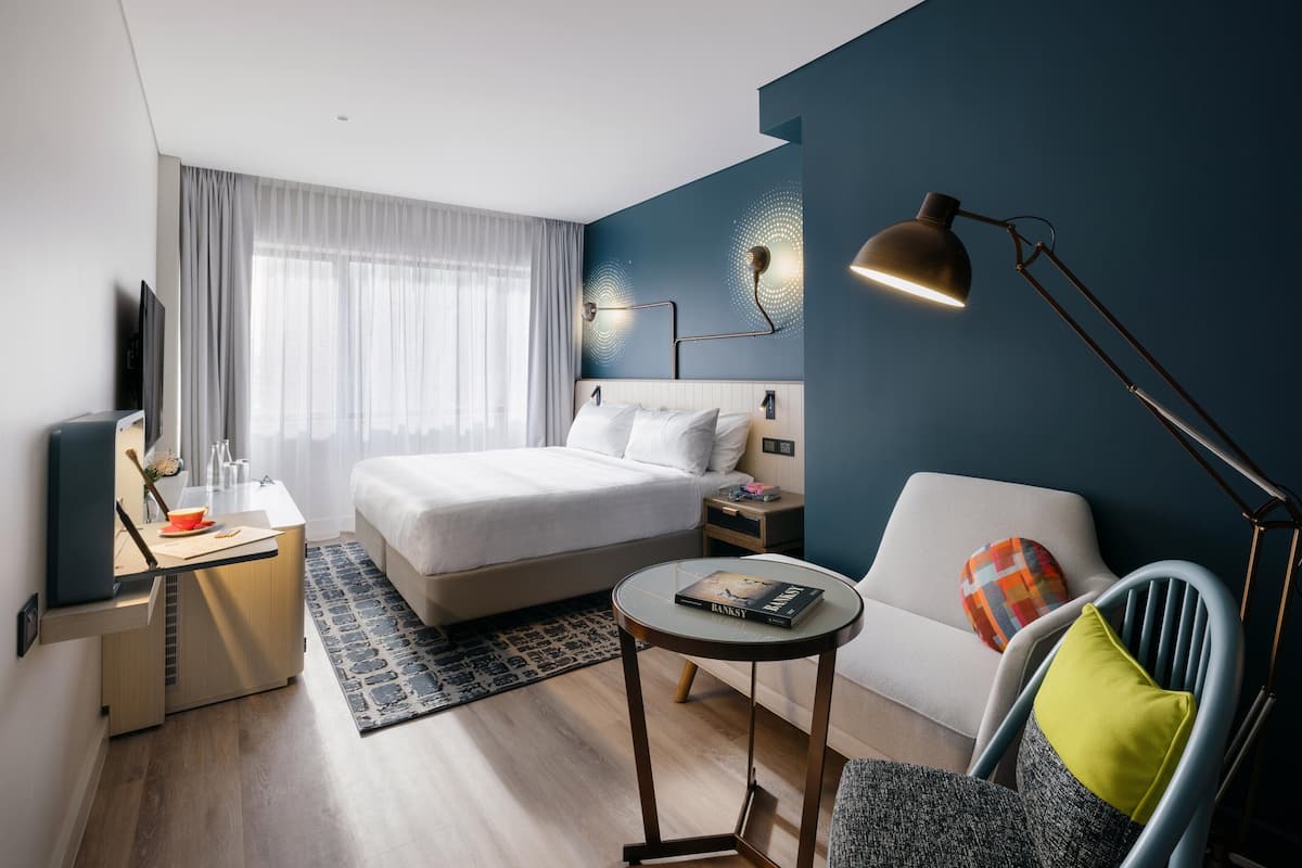 A modern hotel room with a bed, a desk, a chair, a round table, and a TV.