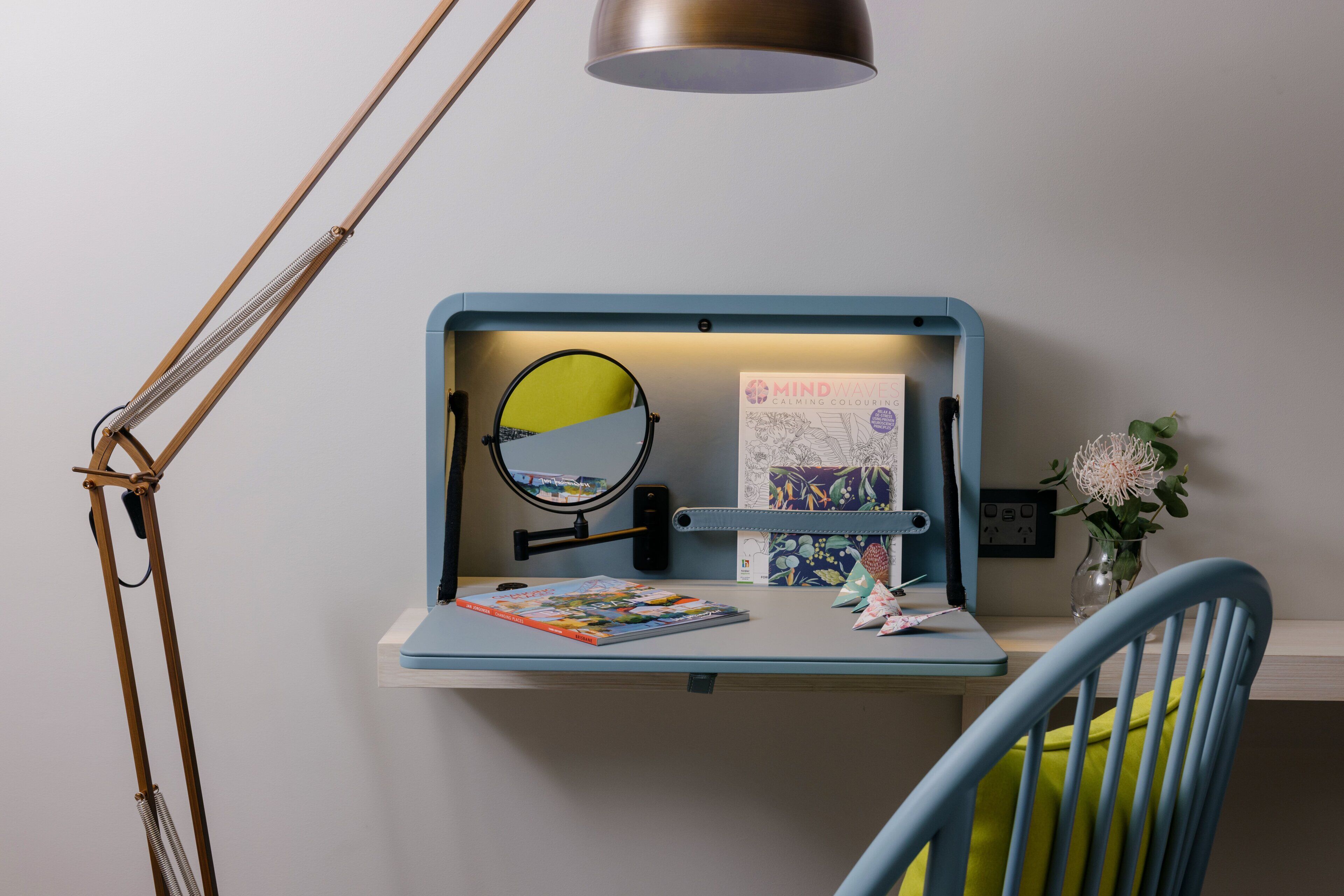 A desk with a lamp, a mirror, a book, and a vase with flowers.