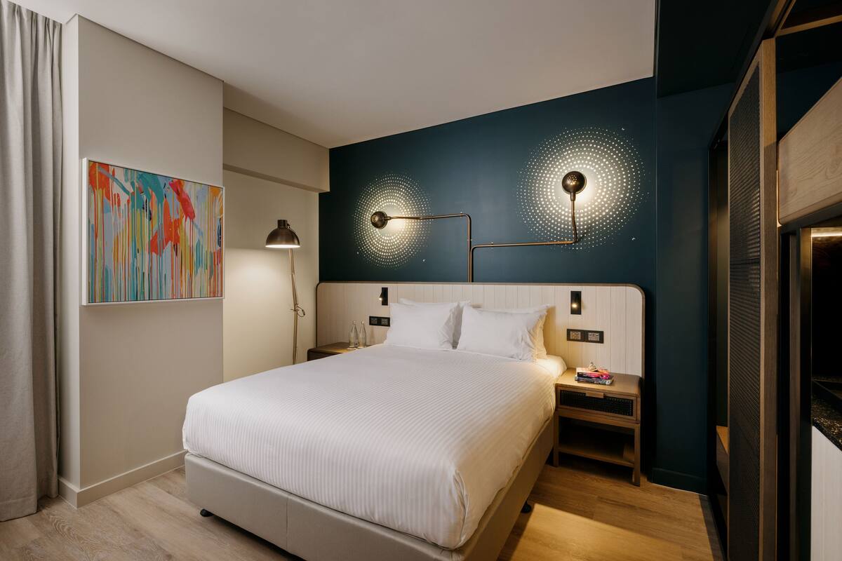 A modern hotel room with a large bed, a desk, and a wall-mounted lamp.