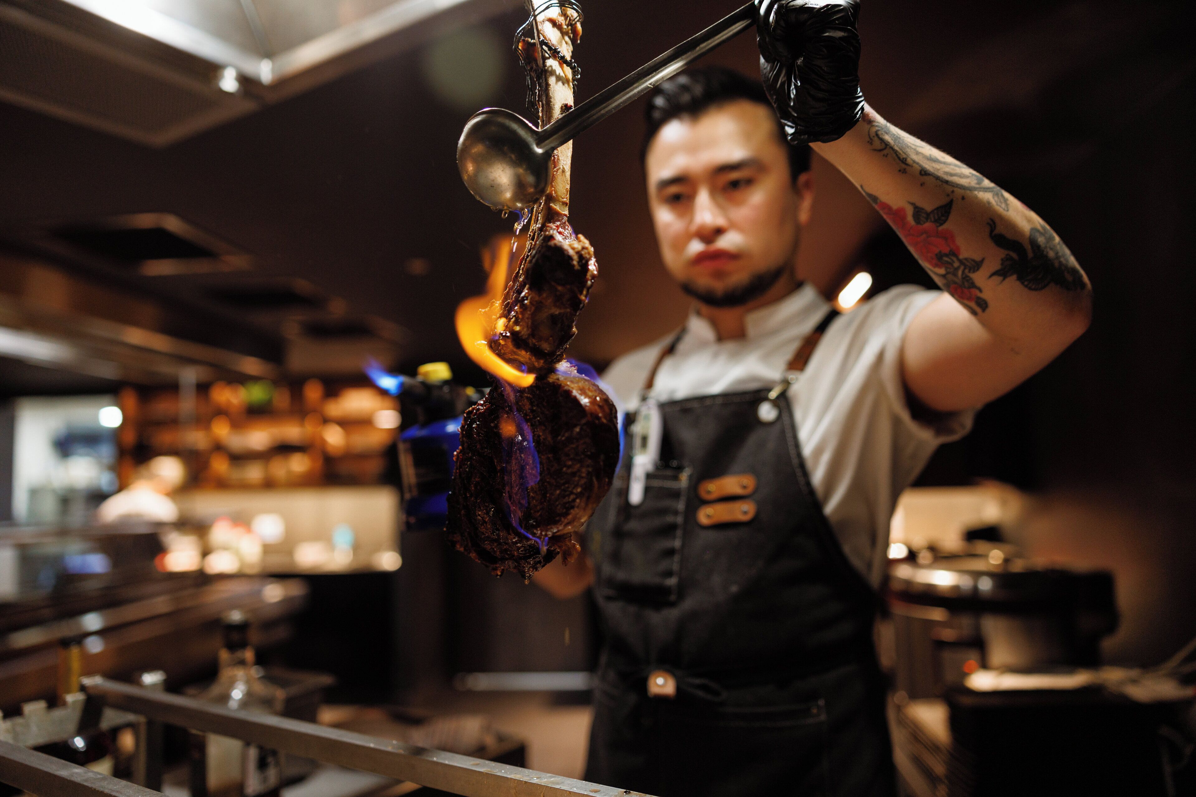 A person in a kitchen setting, wearing a white shirt and black apron, holding a meat with a metal hook.