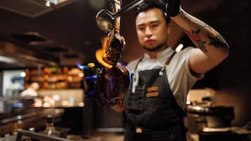 A person in a kitchen setting, wearing a white shirt and black apron, holding a meat with a metal hook.