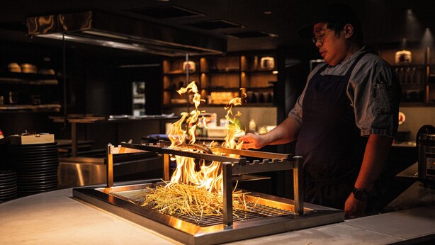 Un chef cocina sobre una parrilla con llamas en una cocina.