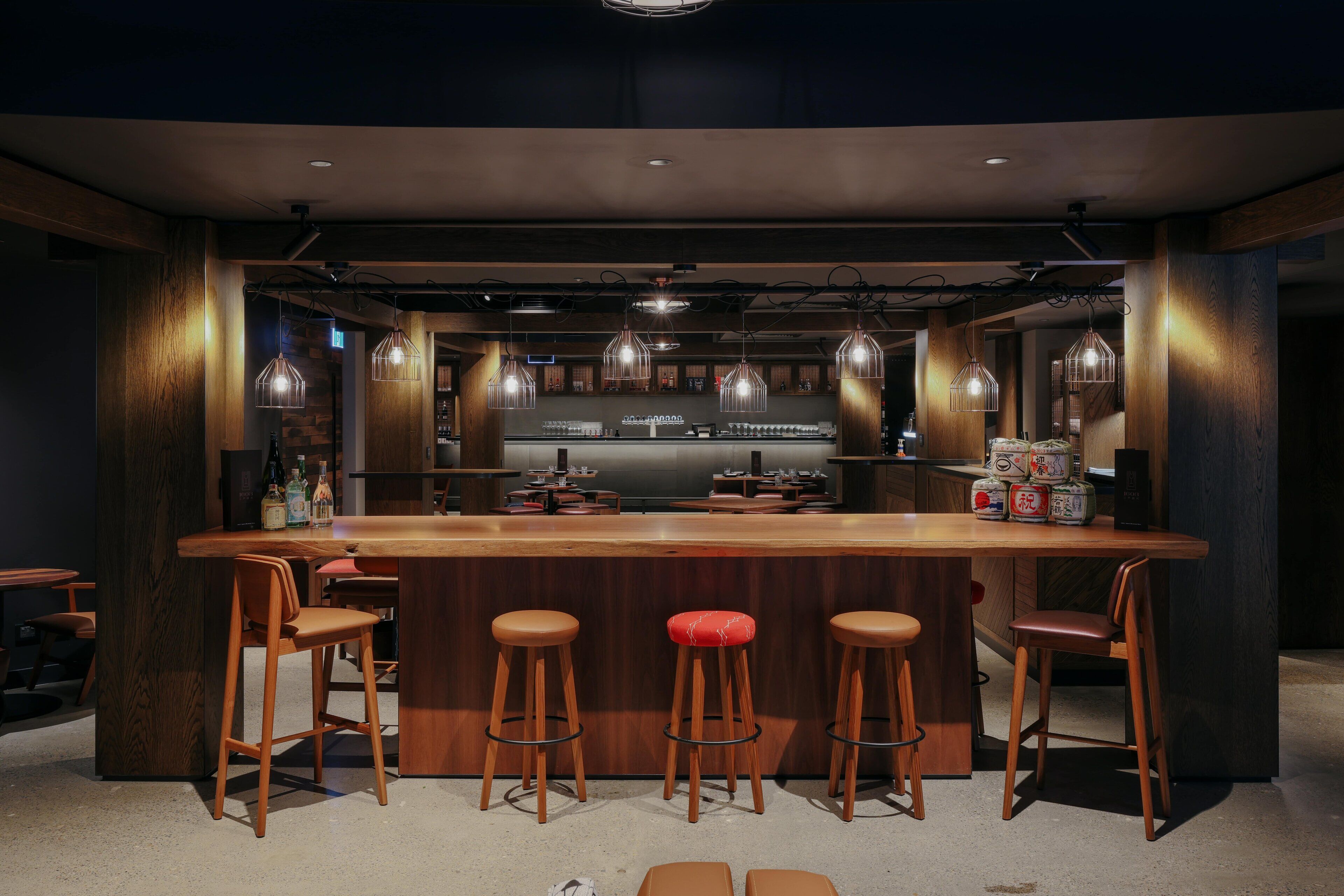 A bar area with a wooden bar counter, stools, hanging pendant lights, and a bar stocked with bottles.