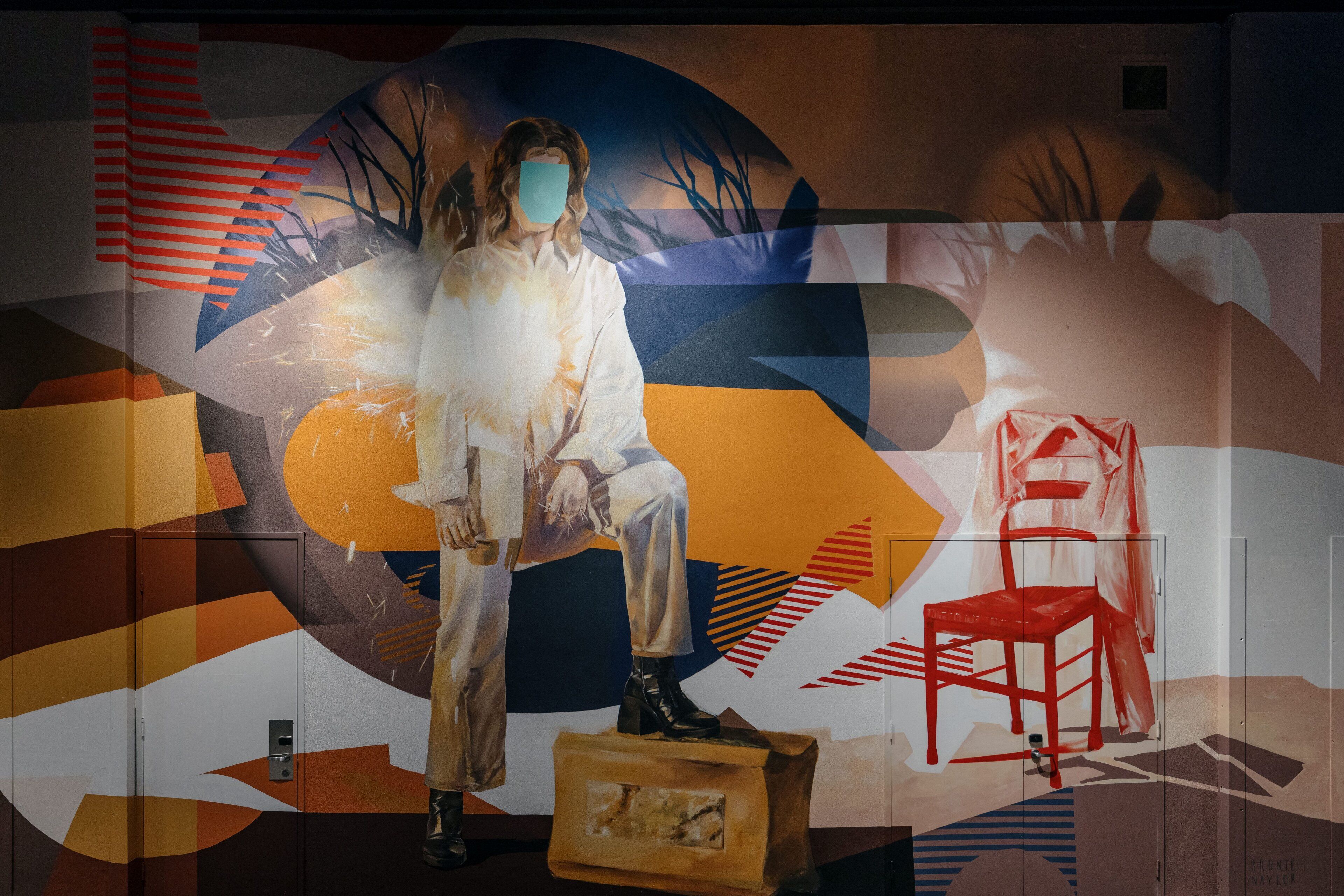 A person sitting on a suitcase in front of a colorful abstract mural.