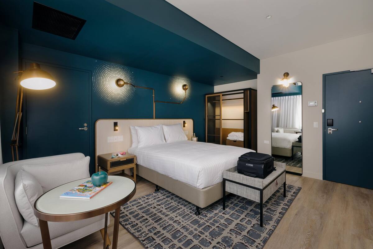 A modern hotel room with a large bed, a sitting area with a small table and chair, a bedside table with a lamp, and a large mirror.