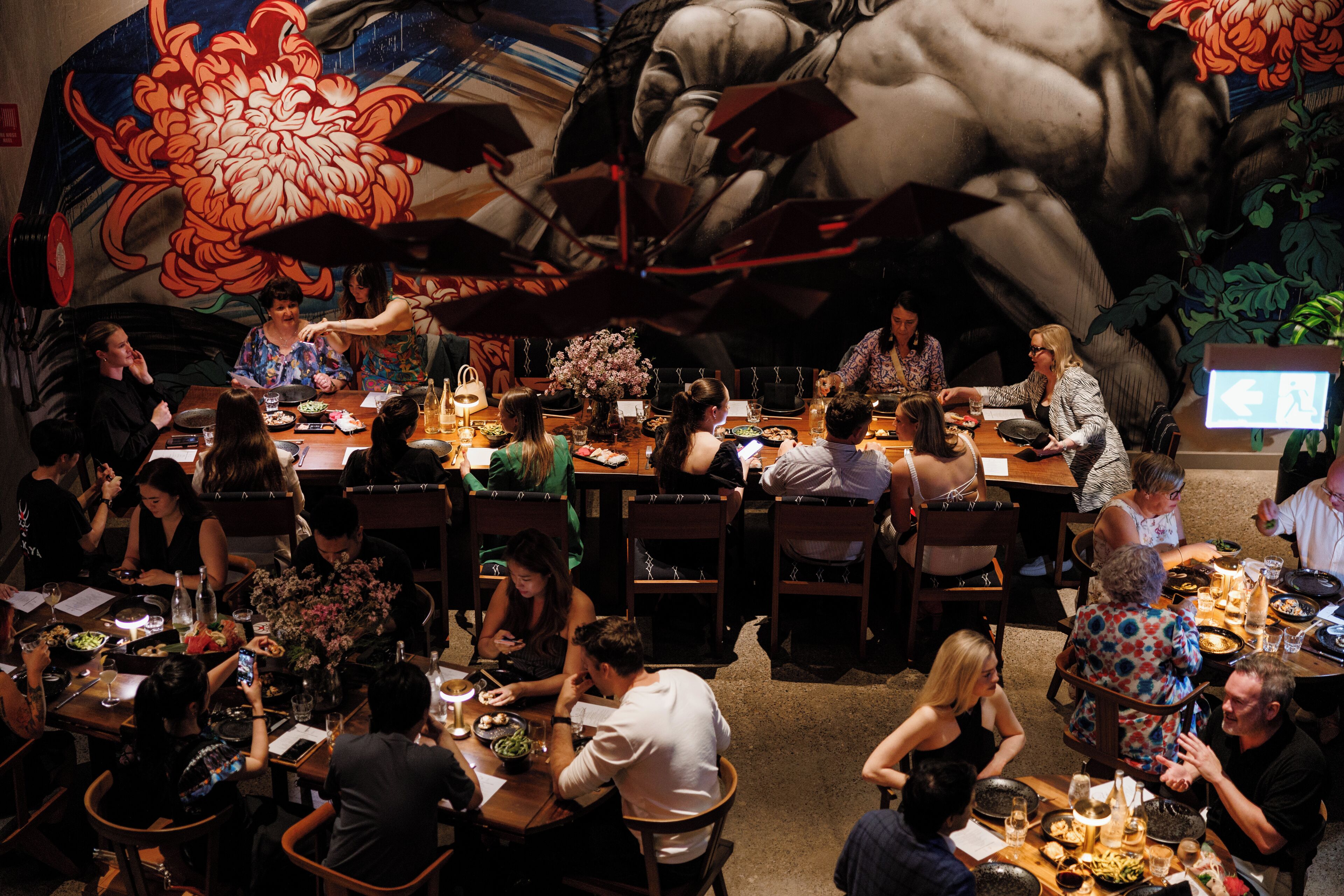 A restaurant with multiple dining tables and chairs, a mural on the wall, and a variety of dishes and drinks on the tables.