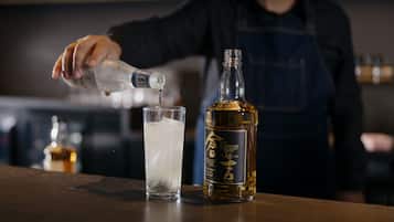 A person pouring a clear liquid into a glass with ice. A bottle labeled "The Kuroshio Whisky" is on the table.