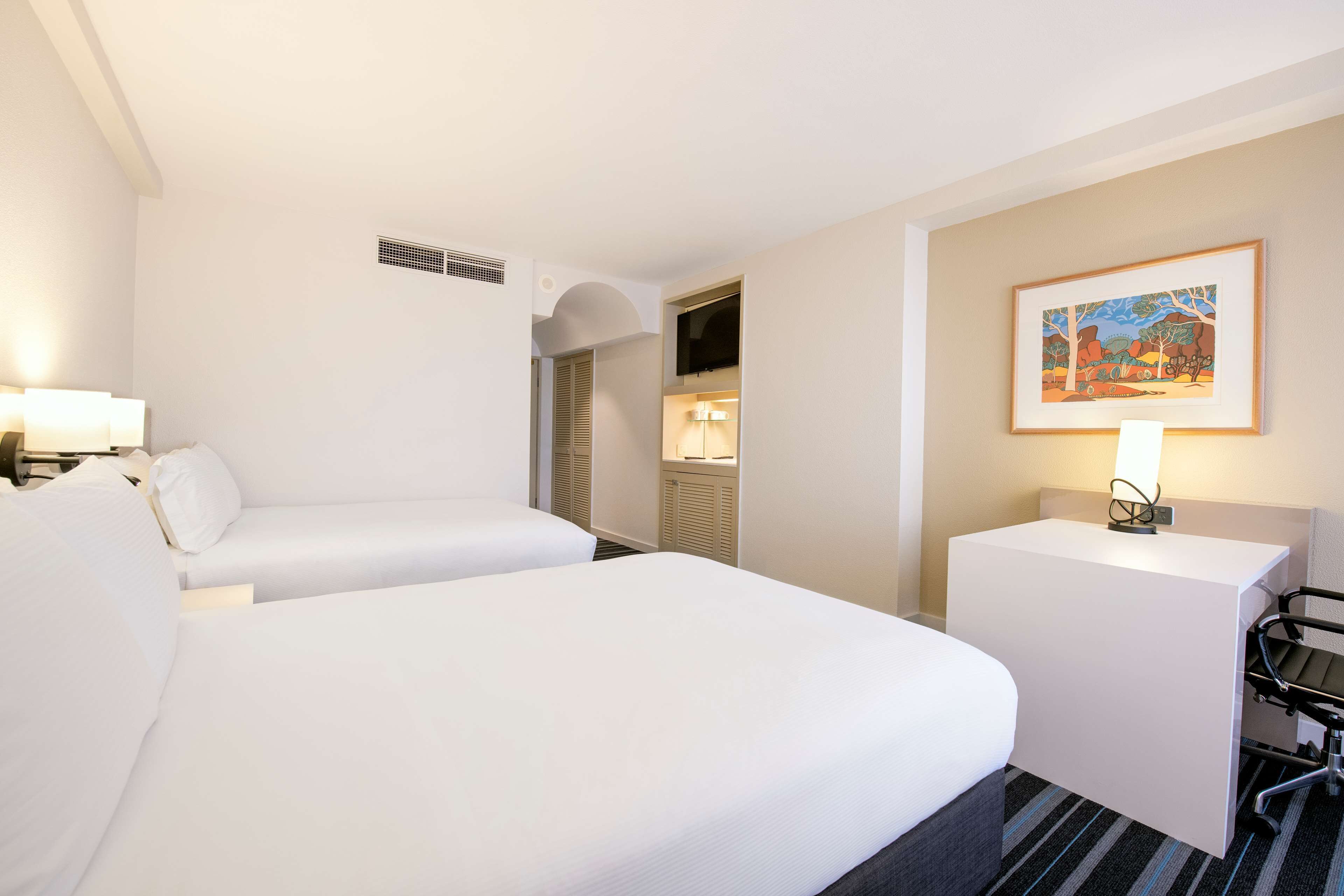 Quarto de hotel com duas camas, uma escrivaninha, uma cadeira, um quadro na parede e um abajur.