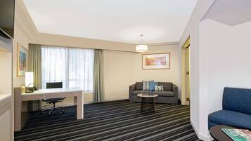 A hotel room with a desk, a sofa, a blue armchair, a patterned carpet, and a painting on the wall.