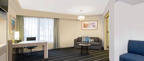Habitación de hotel con escritorio, sofá, sillón azul, alfombra estampada y un cuadro en la pared.