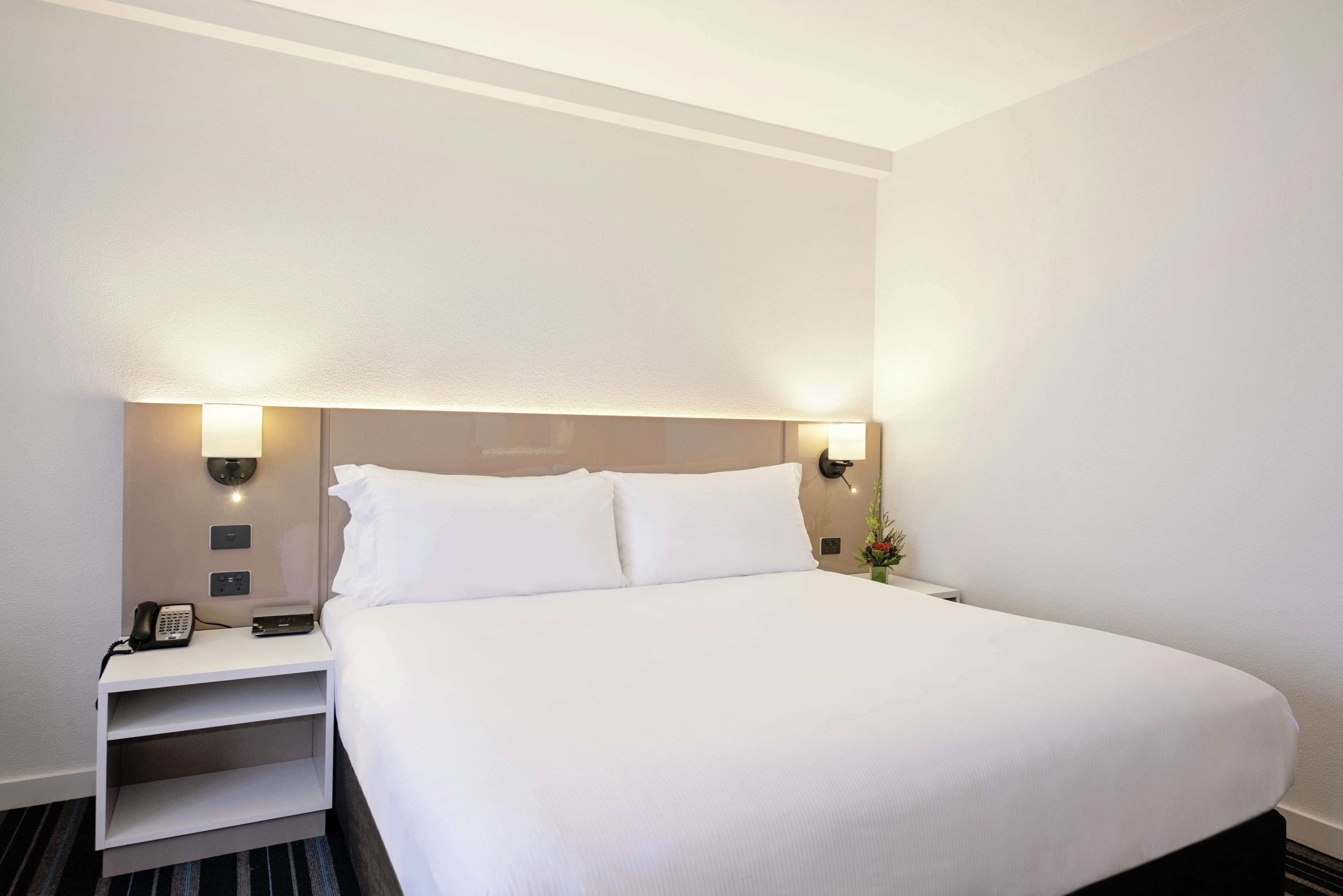 Habitación de hotel con una cama grande, mesitas de noche, un teléfono e iluminación en la pared.