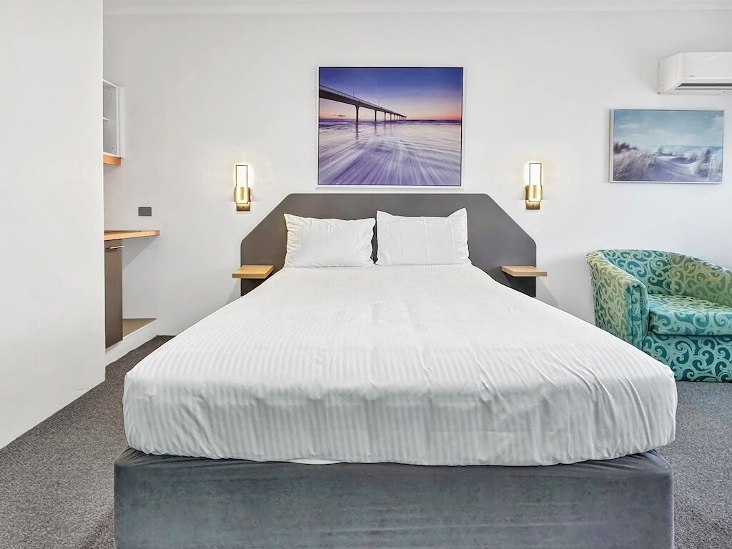 A modern hotel room with a large bed, two bedside tables, a green patterned armchair, and two wall-mounted light fixtures.