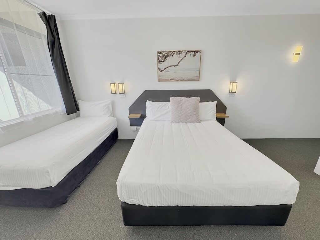 Une chambre d’hôtel avec un grand lit, un plus petit lit, deux lampes de chevet et une fenêtre avec des rideaux légers.