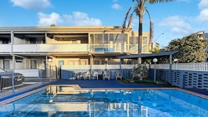 A swimming pool area with lounge chairs and a fence, surrounded by a building with balconies and a palm tree.