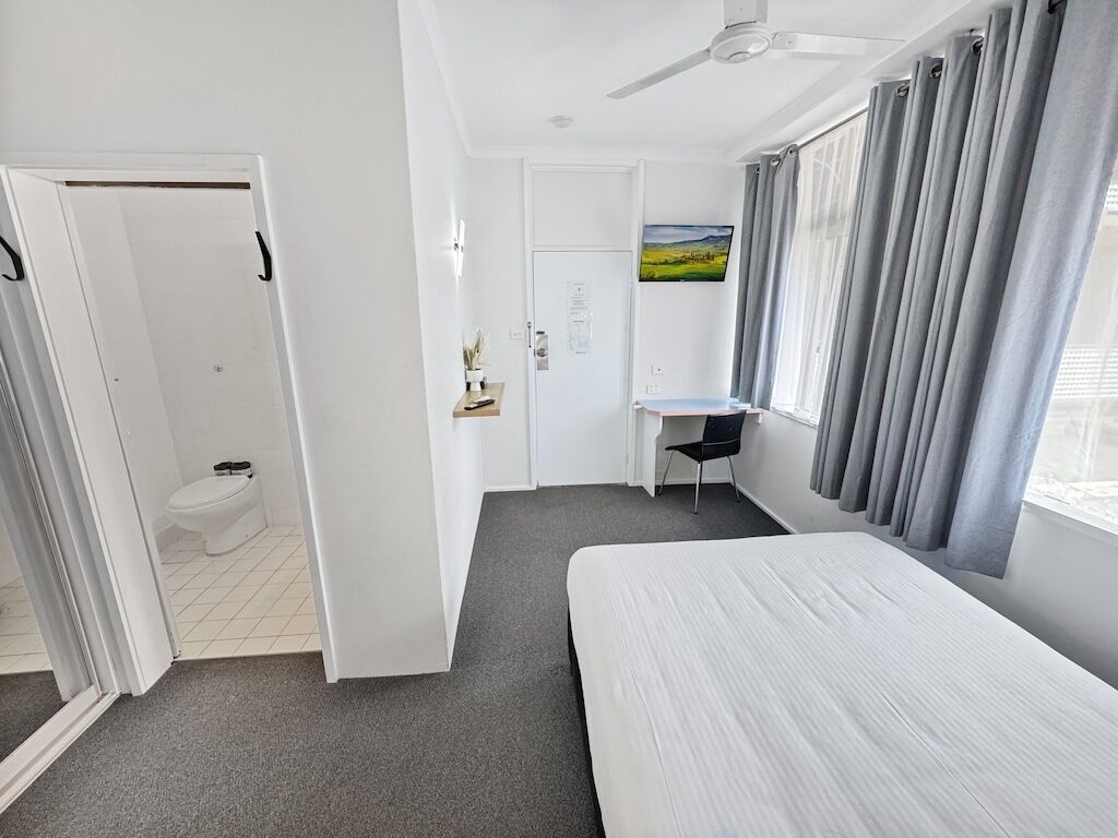 A hotel room with a bed, a desk, a chair, a ceiling fan, and a view of the outdoors.
