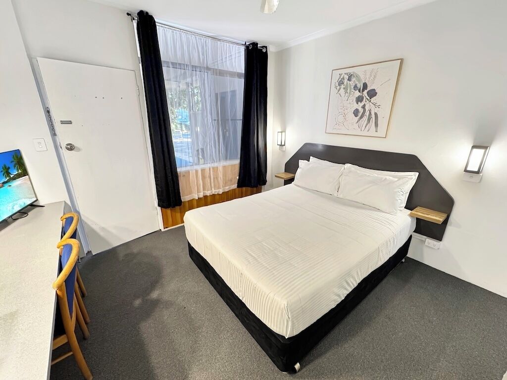 Une chambre à coucher comprenant un lit, un bureau, une chaise, une fenêtre avec des rideaux, un luminaire fixé au mur et un tableau encadré.