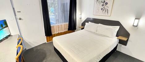 A bedroom with a bed, a desk, a chair, a window with curtains, a wall-mounted light, and a framed picture.